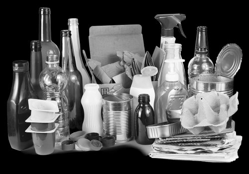 A black-and-white image showing a variety of waste materials collected for rubbish removal, including empty glass bottles with different shapes and sizes, some with narrow necks and others with wider bodies, conveying a mix of beverage containers on a flat surface. There are also aluminum cans, stacked neatly, alongside paper waste such as newspapers or magazines, which are folded and slightly crumpled. A cardboard box partially filled with similar waste is positioned towards the back, with the opening facing the viewer, revealing more paper and packaging materials inside. Additionally, plastic spray bottles and containers are visible, some with spray nozzles attached, indicating cleaning or household liquids. The overall scene appears to be set on a driveway or paved area, with a neutral background emphasizing the collection of rubbish ready for removal by a professional service like House Clearance Byfleet, serving the local area around Guildford postcode GU23. The image represents typical household waste ready for disposal or recycling as part of a rubbish clearance service in the Surrey region.