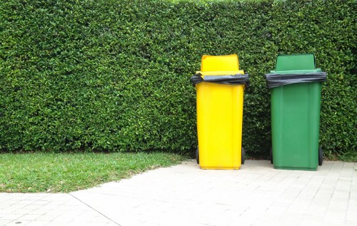 In the image, two large upright wheelie bins are positioned on a paved pathway in front of a neatly trimmed green hedge. The bin on the left is bright yellow with a black lid, while the bin on the right is green with a matching black lid. Each bin appears to be made of durable plastic with a textured surface, typical of waste containers used for household rubbish collection. The yellow bin has some black garbage bags placed loosely on top, leaning slightly to one side, while the green bin does not have bags on top. The surrounding environment suggests an outdoor setting, possibly a driveway or garden area adjacent to a residential property in a suburban area, with natural daylight illuminating the scene. The image relates to rubbish collection or waste management services, such as those offered by House Clearance Byfleet, emphasizing general waste disposal tasks. The scene is clean, orderly, and highlights the practical use of waste bins for household rubbish during routine clearance or disposal activities in the local area.