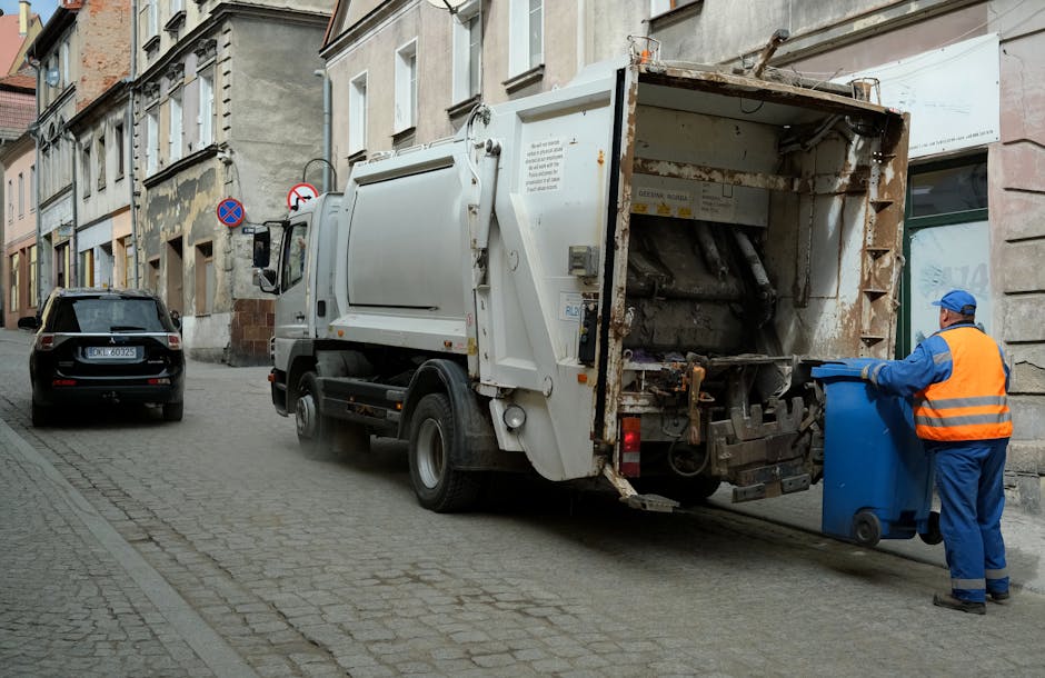 A sanitation worker dressed in a blue uniform with a bright orange reflective vest and a matching blue cap is positioned on the right side of the image, handling a blue wheeled rubbish bin with both h