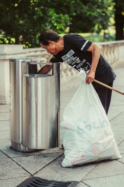A sanitation worker dressed in a blue uniform with a bright orange reflective vest and a matching blue cap is positioned on the right side of the image, handling a blue wheeled rubbish bin with both h