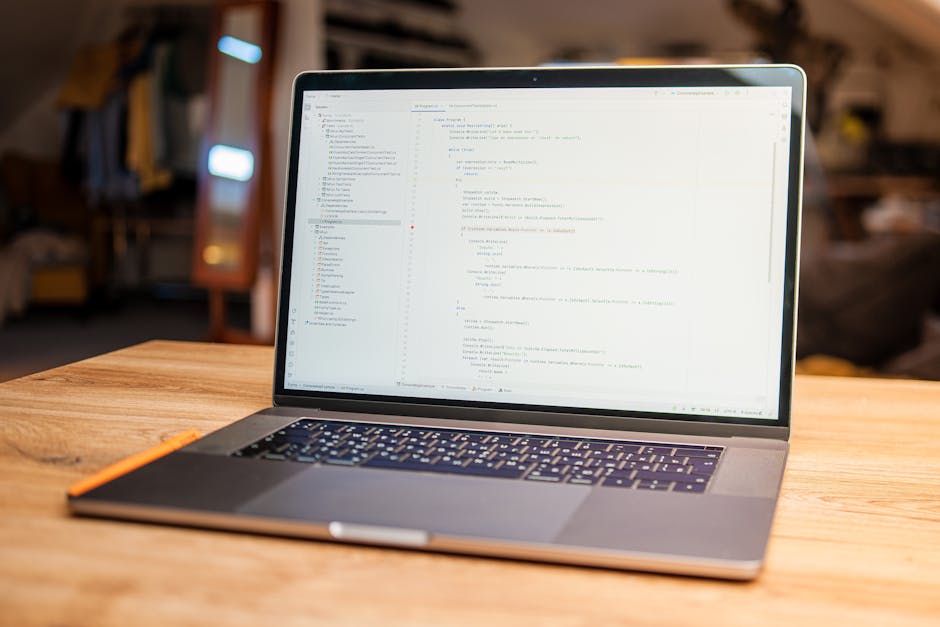 A modern, open-plan workspace featuring a silver laptop placed on a smooth wooden desk. The laptop's screen displays lines of light-colored code in a text editor, with a visible sidebar and multiple o