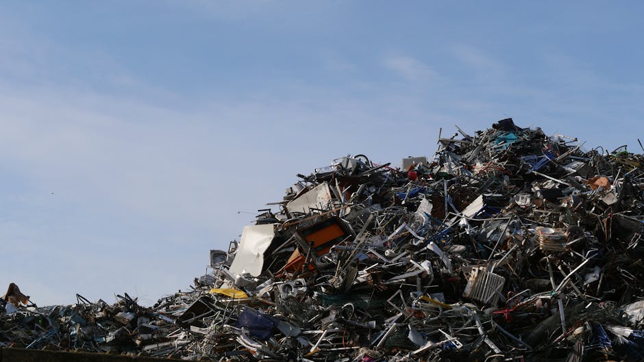 A narrow urban alleyway filled with a large amount of waste and discarded materials, primarily comprising stacked cardboard boxes, black plastic bags, and various bulky rubbish. In the foreground, a l