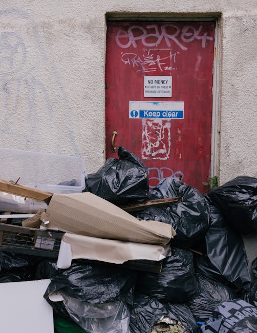 A pile of mixed waste materials including cardboard boxes, paper, and plastic packaging sacks is gathered at the base of a brick wall adjacent to a tree trunk. The cardboard boxes, varying in size and